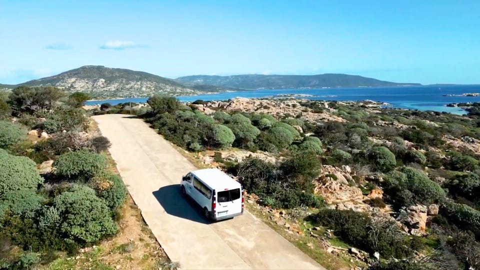 Un minivan bianco su una pista di cemento tra la macchia bassa, con una baia azzurra e colline sullo sfondo