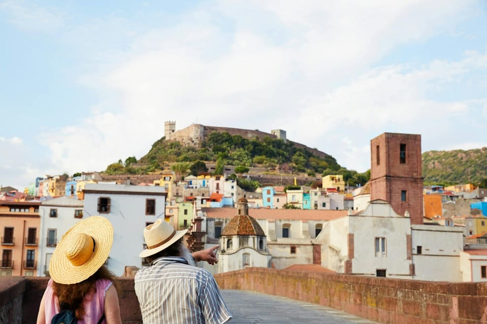 Two visitors in sun hats on a stone bridge looking across at pastel-coloured houses and a hilltop castle