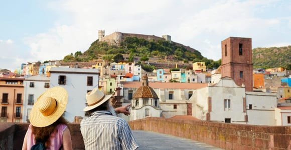 Two visitors in sun hats on a stone bridge looking across at pastel-coloured houses and a hilltop castle