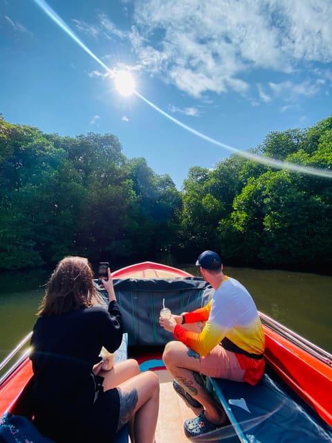 Two passengers sitting in the bow of a narrow red open boat on a mangrove channel, sunlight flaring over the treeline