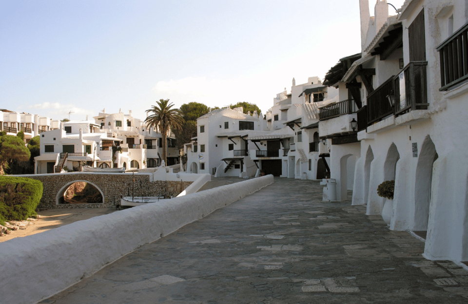 A whitewashed village street with arched doorways and a low white wall, a palm tree and a small stone bridge in the background