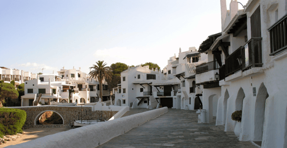 A whitewashed village street with arched doorways and a low white wall, a palm tree and a small stone bridge in the background