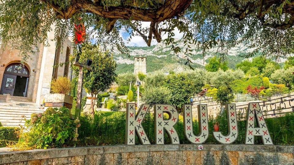 Large lettering spelling Kruja in a garden beneath olive branches, a stone building on the left and a mountainside behind