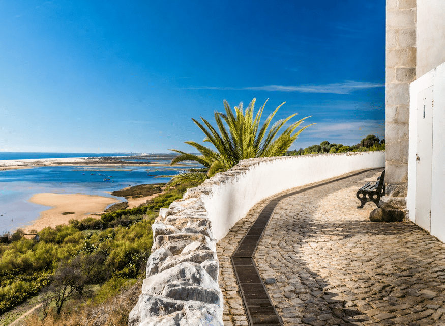A cobbled path curving along a whitewashed wall above the lagoon, with a palm and a bench overlooking sandbanks and blue water