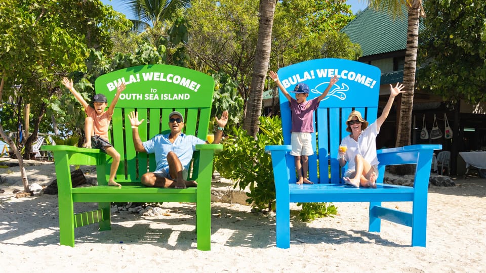 Two adults and two children waving from a pair of oversized green and blue wooden beach chairs painted with BULA! WELCOME!, palms and white sand behind them
