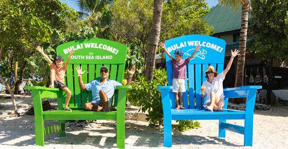 Two adults and two children waving from a pair of oversized green and blue wooden beach chairs painted with BULA! WELCOME!, palms and white sand behind them