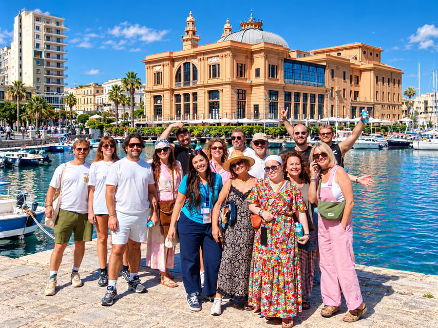 A group of smiling visitors posing on the harbour quay in Bari, with a domed historic building and moored boats behind them