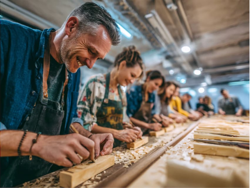 A row of people in work aprons bent over a long wooden bench, each shaping a pale block of wood with a hand tool among heaps of shavings