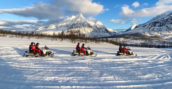 Three snowmobiles with riders in red suits stopped on a snowfield, a pointed snow-covered peak behind them under a blue sky