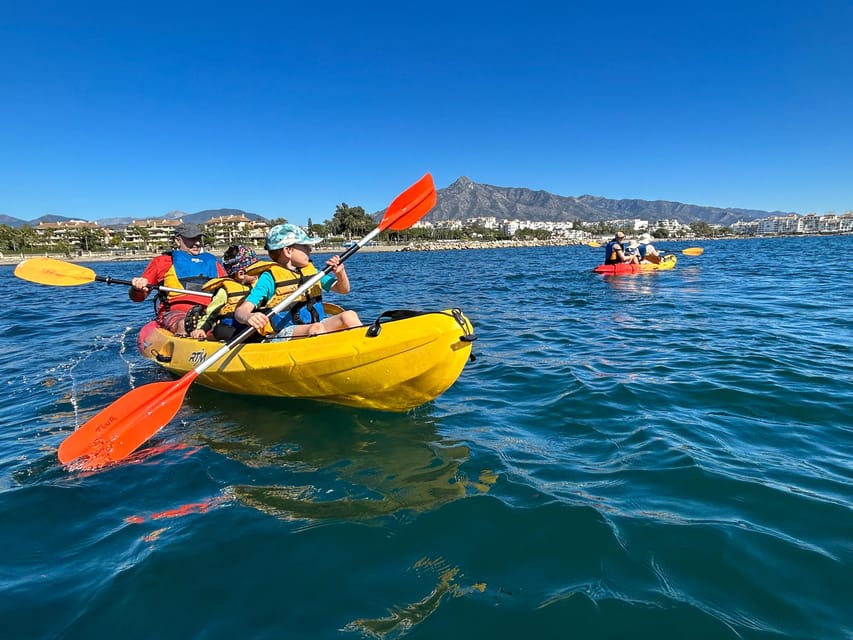 An adult and two children in life jackets paddling a yellow kayak on blue water with a mountain behind the coast
