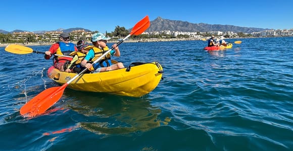 An adult and two children in life jackets paddling a yellow kayak on blue water with a mountain behind the coast