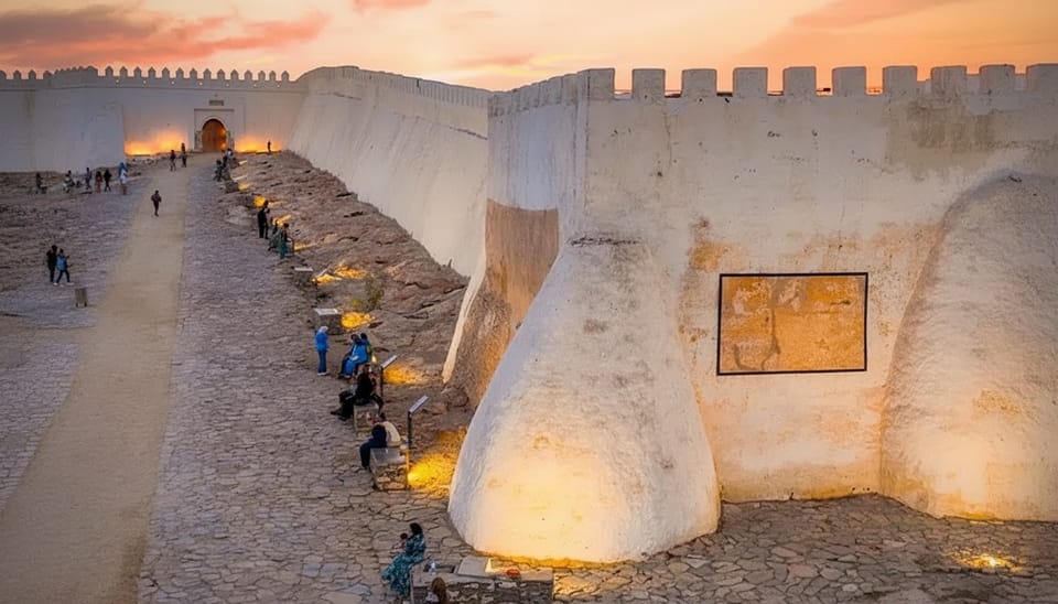 Whitewashed hilltop fortress walls with a lit arched gateway at dusk, visitors sitting on benches along the sloping path beside the rampart
