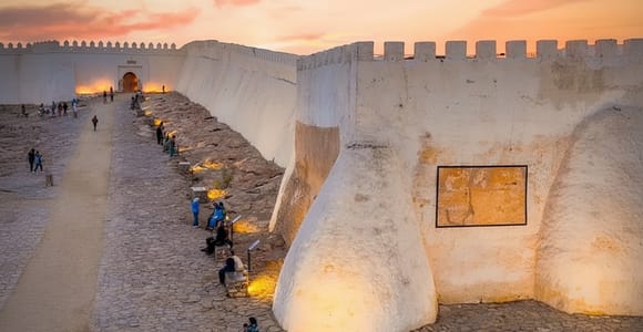 Whitewashed hilltop fortress walls with a lit arched gateway at dusk, visitors sitting on benches along the sloping path beside the rampart