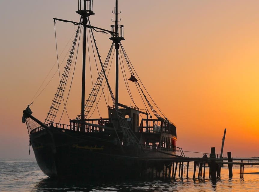 A dark wooden pirate ship with two masts moored at a jetty against an orange sunset sky