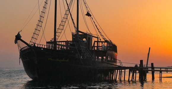 A dark wooden pirate ship with two masts moored at a jetty against an orange sunset sky