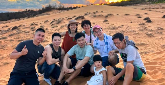 A group of travellers sitting with a small child in white on a sand ridge under an orange sunset sky