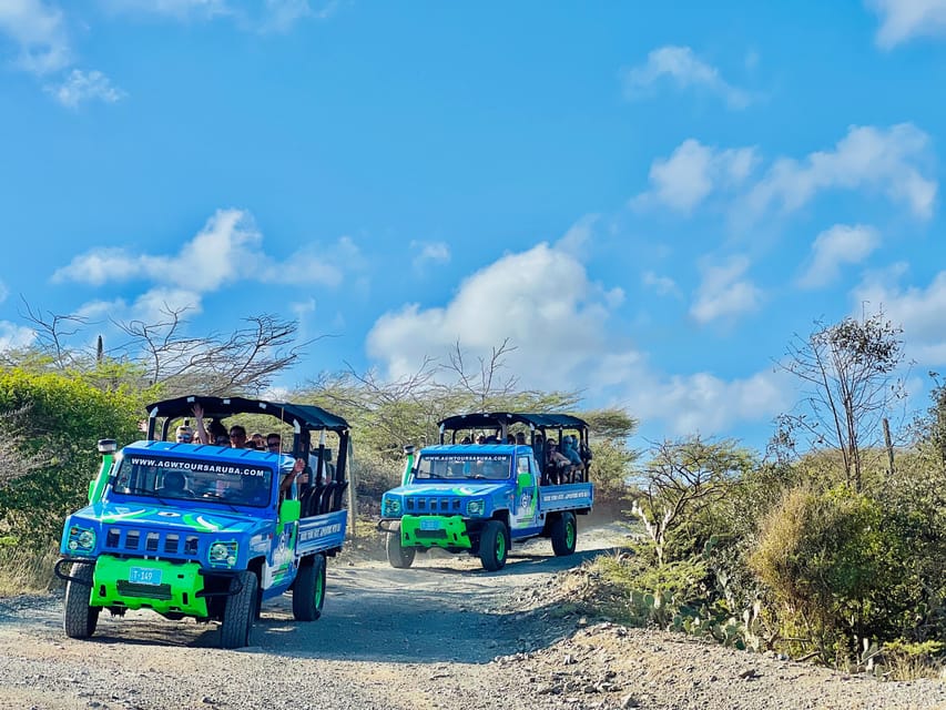 Two open blue-and-green off-road trucks with passengers on a gravel track between scrub