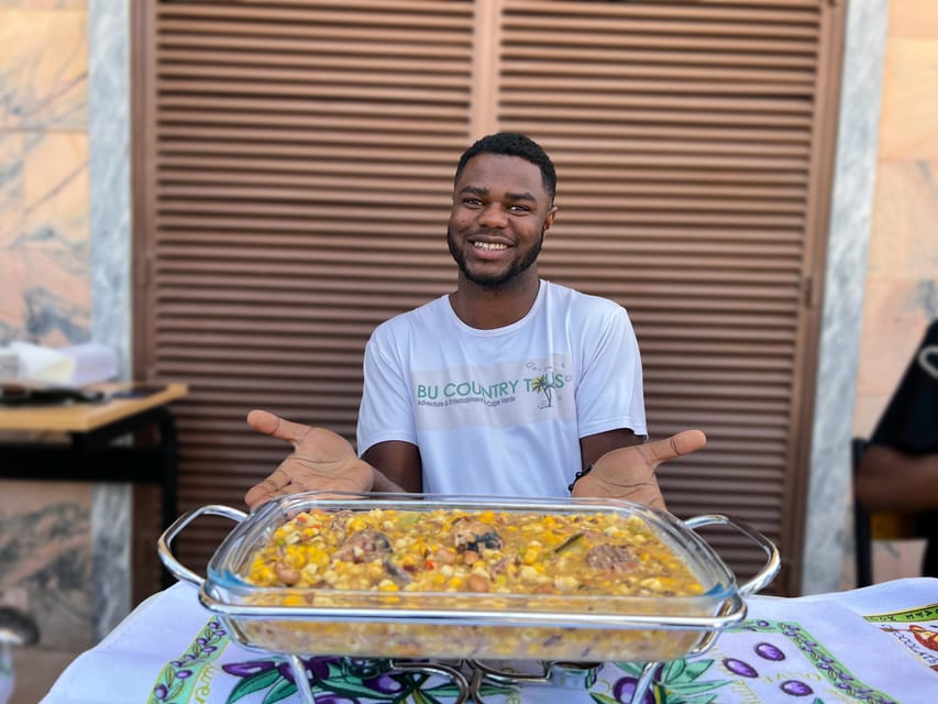A young man in a tour company T-shirt holding out a glass dish of maize and bean stew on a patterned tablecloth