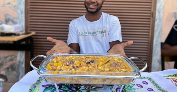 A young man in a tour company T-shirt holding out a glass dish of maize and bean stew on a patterned tablecloth