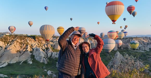 A couple making a heart shape with their arms in front of dozens of hot-air balloons above a rocky valley at sunrise