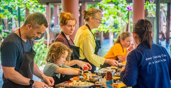 A cooking class in progress: participants in aprons at a long worktop with portable gas burners, the host in a branded polo shirt, greenery beyond the open side of the room
