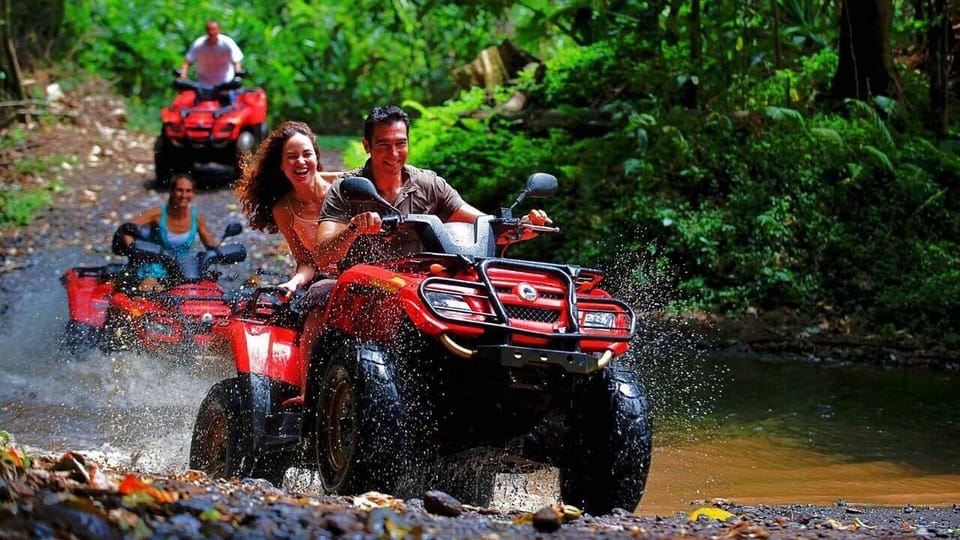 Two muddy quad bikes with riders in helmets crossing a shallow stream in a pine forest