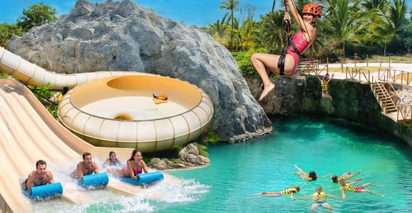 Water park with a curving slide running into a turquoise pool, a person on a zip line overhead and children floating in yellow life jackets below