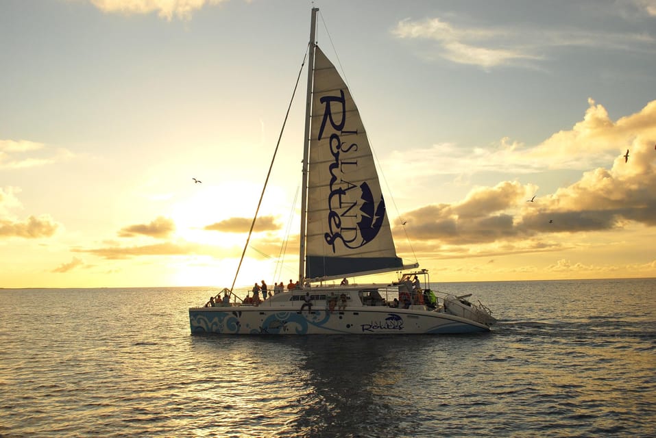 A catamaran under full sail on a calm sea at dusk, passengers lining the deck, the sky behind it gold and grey with low cloud