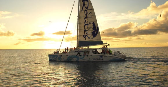 A catamaran under full sail on a calm sea at dusk, passengers lining the deck, the sky behind it gold and grey with low cloud
