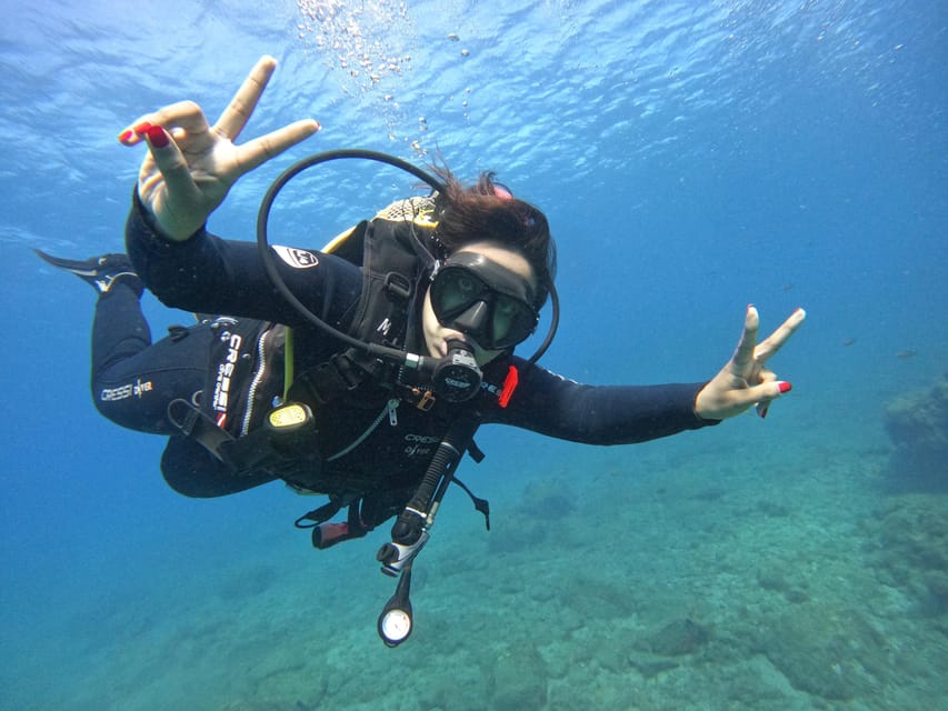 A scuba diver in a black wetsuit and mask swimming in clear blue water above a rocky seabed, making a peace sign with both hands toward the camera