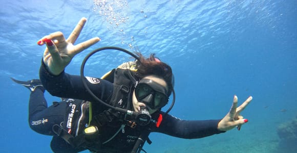 A scuba diver in a black wetsuit and mask swimming in clear blue water above a rocky seabed, making a peace sign with both hands toward the camera