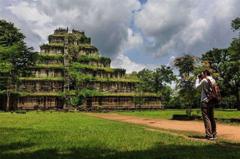 Die steinerne Stufenpyramide des Prasat Thom in Koh Ker über dem Wald, davor Rasen und ein Sandweg, ein Besucher fotografiert sie