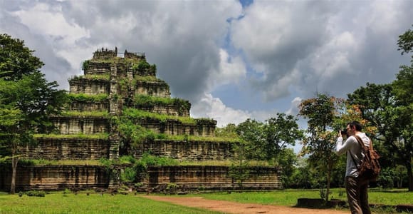 The stepped stone pyramid of Prasat Thom at Koh Ker rising above the forest, grass and a dirt path in front, a visitor photographing it