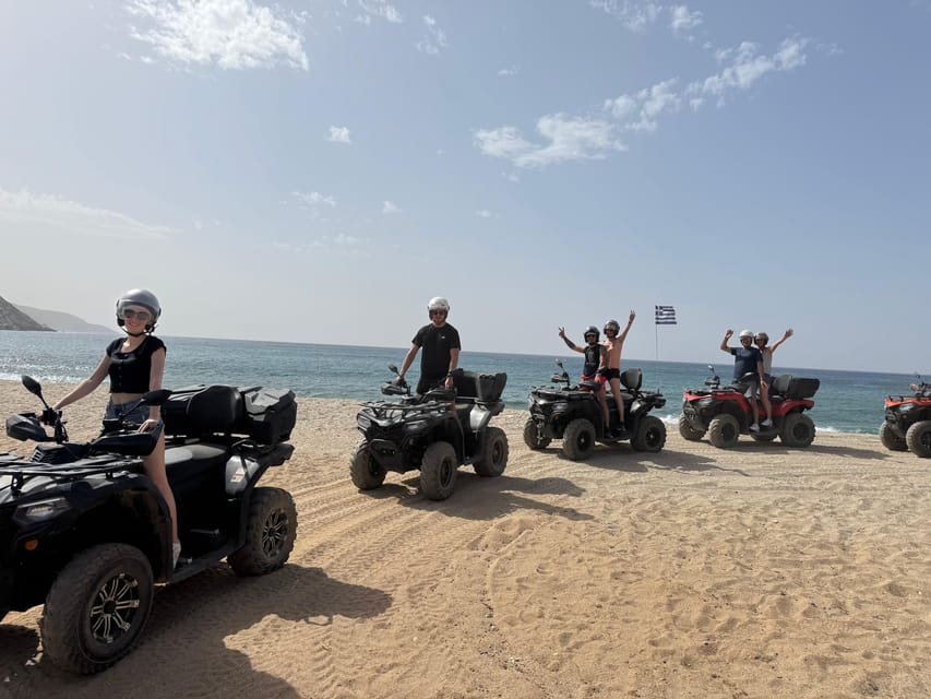 Black and red quad bikes parked on a sandy beach with the sea behind, riders in helmets waving and a Greek flag on a pole
