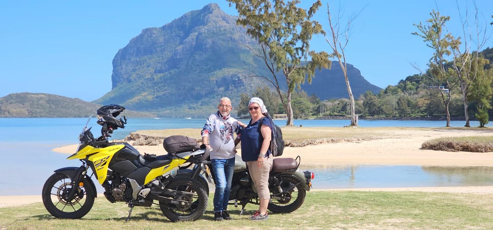Two hired motorbikes parked on grass beside a shallow turquoise lagoon, a couple standing between them, with the dark bulk of Le Morne Brabant rising behind