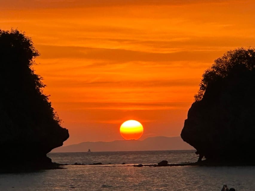 Longtail boat silhouetted on flat water in front of limestone islands as the sky turns orange