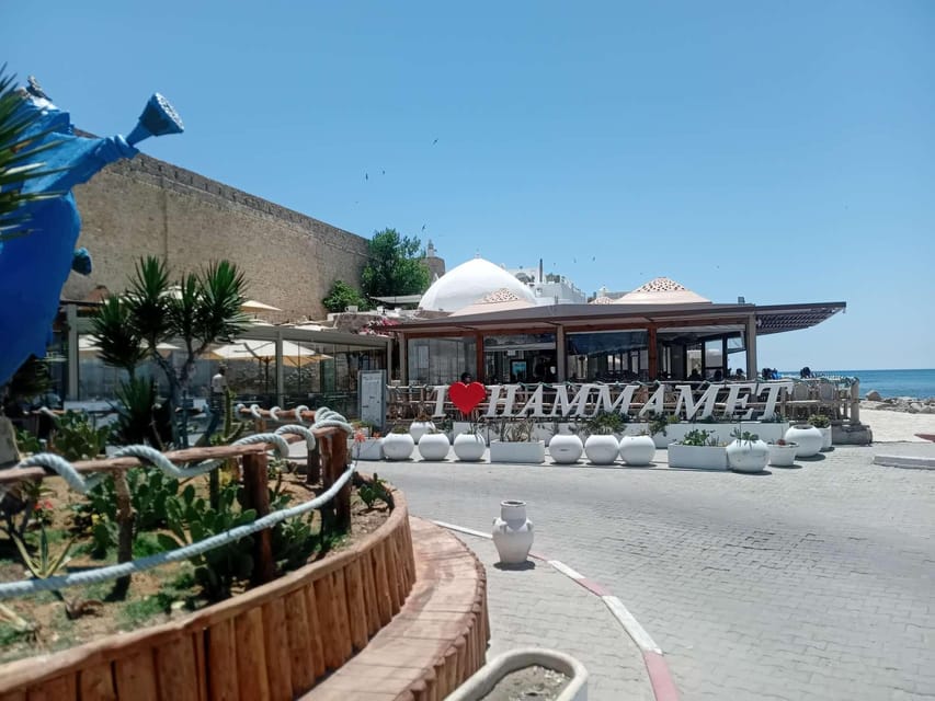 Sand-coloured medina ramparts beside a seafront café with an "I love Hammamet" sign, white domes and the open sea behind