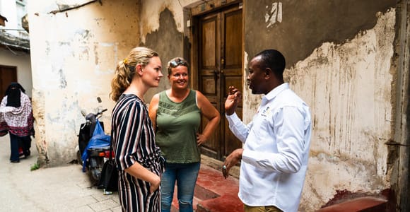 A guide in a white shirt talking to two visitors in a narrow alley, a weathered carved door behind them