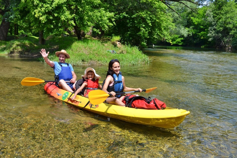 A woman, a man and a child in life jackets sit in a yellow kayak on shallow, clear river water