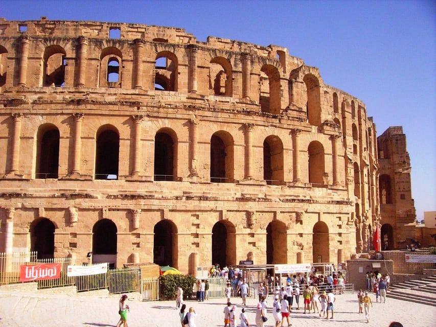 The tiered stone arcades of the El Jem amphitheatre in warm sunlight, with a crowd of visitors in the open square below