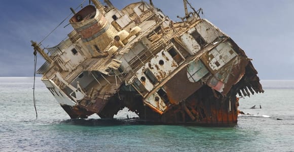 A rusting cargo ship lying grounded and heavily listing on a shallow reef in open water, its white superstructure streaked with rust and its stern broken open, under a pale blue sky