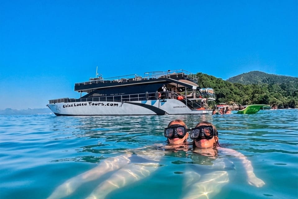 Two snorkellers in masks surfacing in clear blue water in front of a large black-and-white catamaran anchored off a green headland