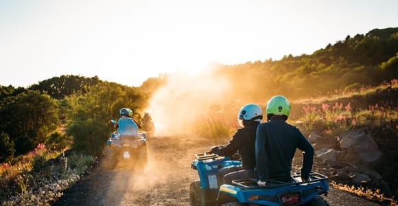 Two riders in helmets on a blue quad bike on a dusty track in low golden light, a second quad behind
