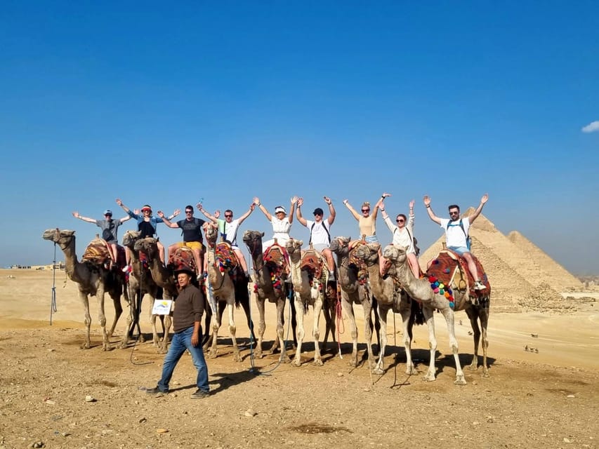 A group of travellers sitting on decorated camels in a row in front of two of the Giza pyramids, arms raised, with a handler standing in the sand in front of them