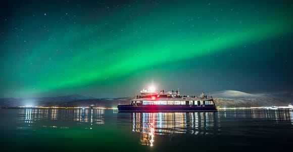A lit passenger boat sits on dark calm water beneath a green aurora band, with city lights along the far shore