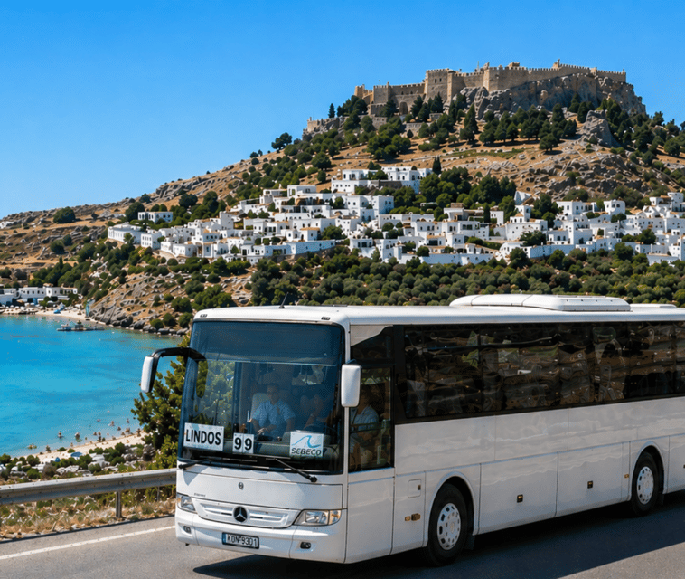 A white coach marked Lindos on a coast road, with the whitewashed village on the hillside and the fortress walls on the crest above a blue bay
