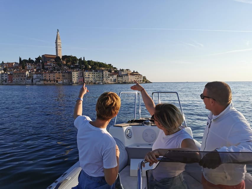 Three people on the front of a small speedboat pointing towards Rovinj’s old town and church tower across calm water