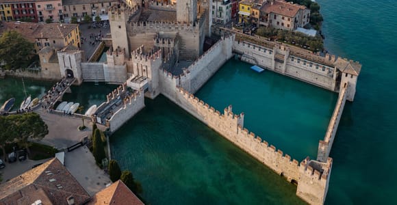 Aerial view of Sirmione's moated Scaliger Castle beside turquoise lake water, with a small white boat below