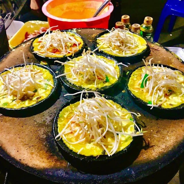 Small cast-iron pans set into a round griddle at a night stall, each holding a turmeric-yellow pancake heaped with raw bean sprouts