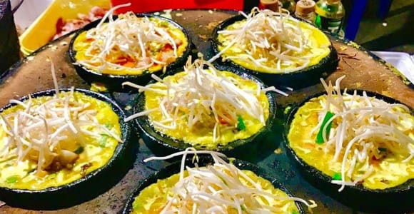 Small cast-iron pans set into a round griddle at a night stall, each holding a turmeric-yellow pancake heaped with raw bean sprouts